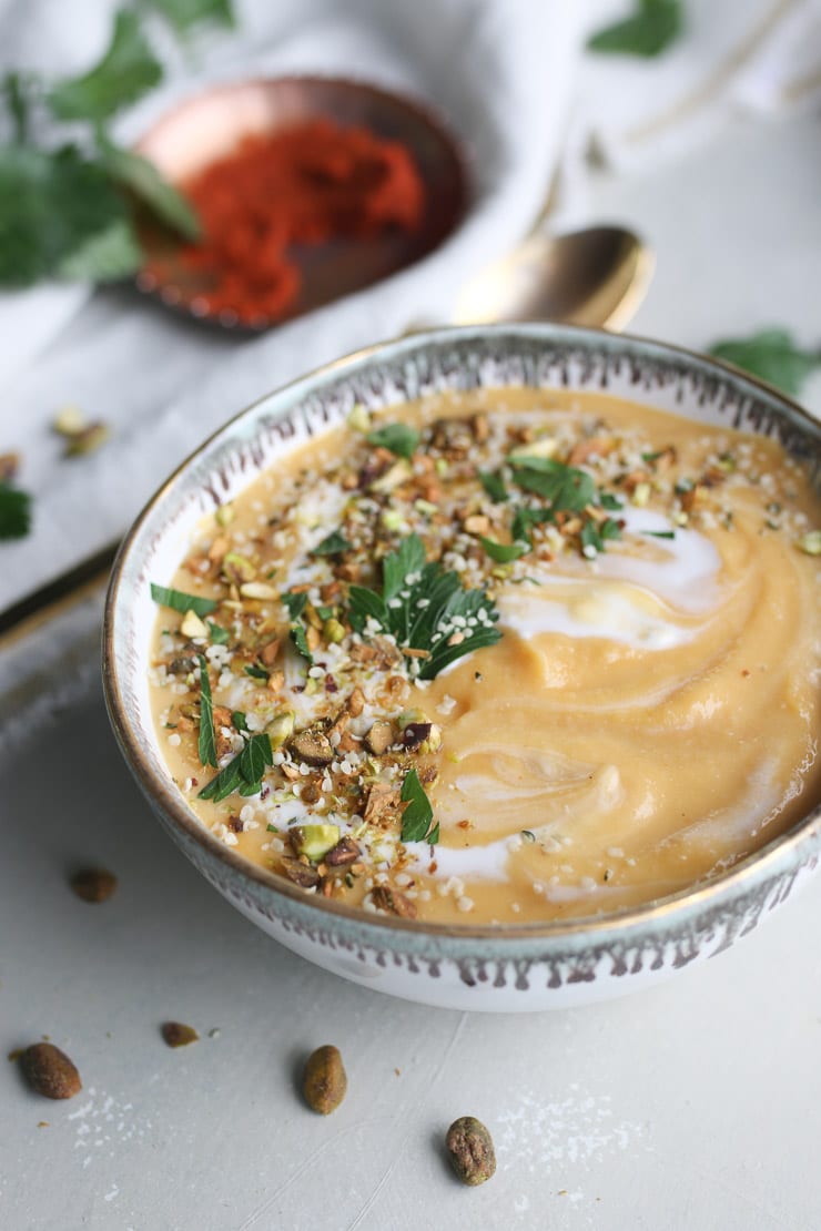 Image of lentil butternut squash soup in a white bowl topped with parsley and nuts with red curry paste in the background.