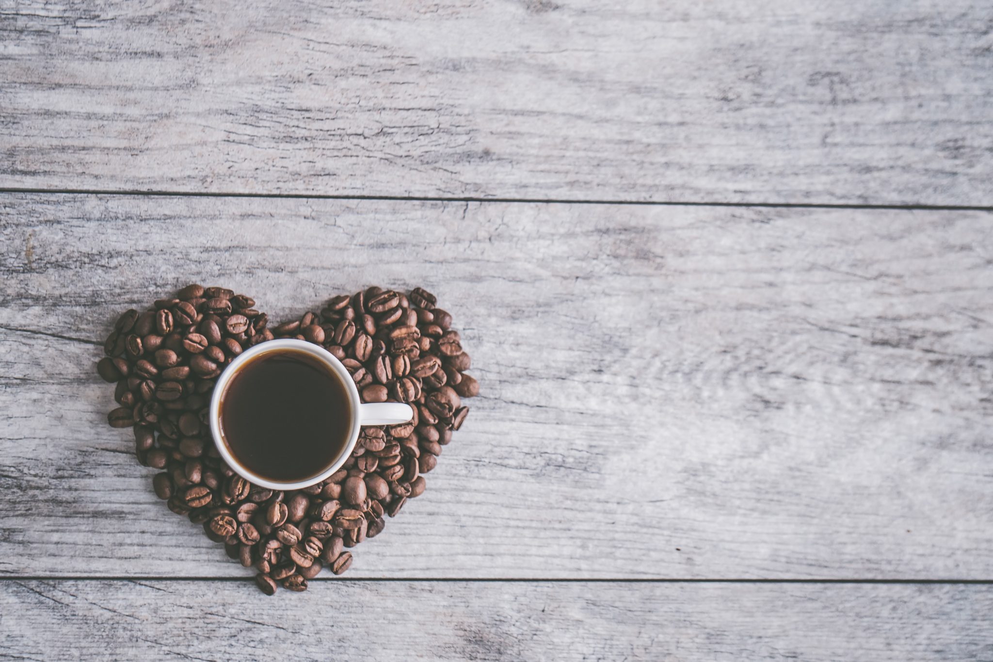 birds eye view of coffee beans in a heart shape with a mug of coffee in the centre against a wooden background