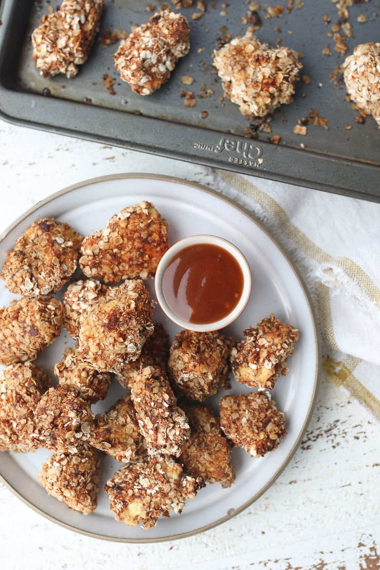 Birds eye view of tofu nuggets on a white plate with a dipping sauce with baking sheet in the background.