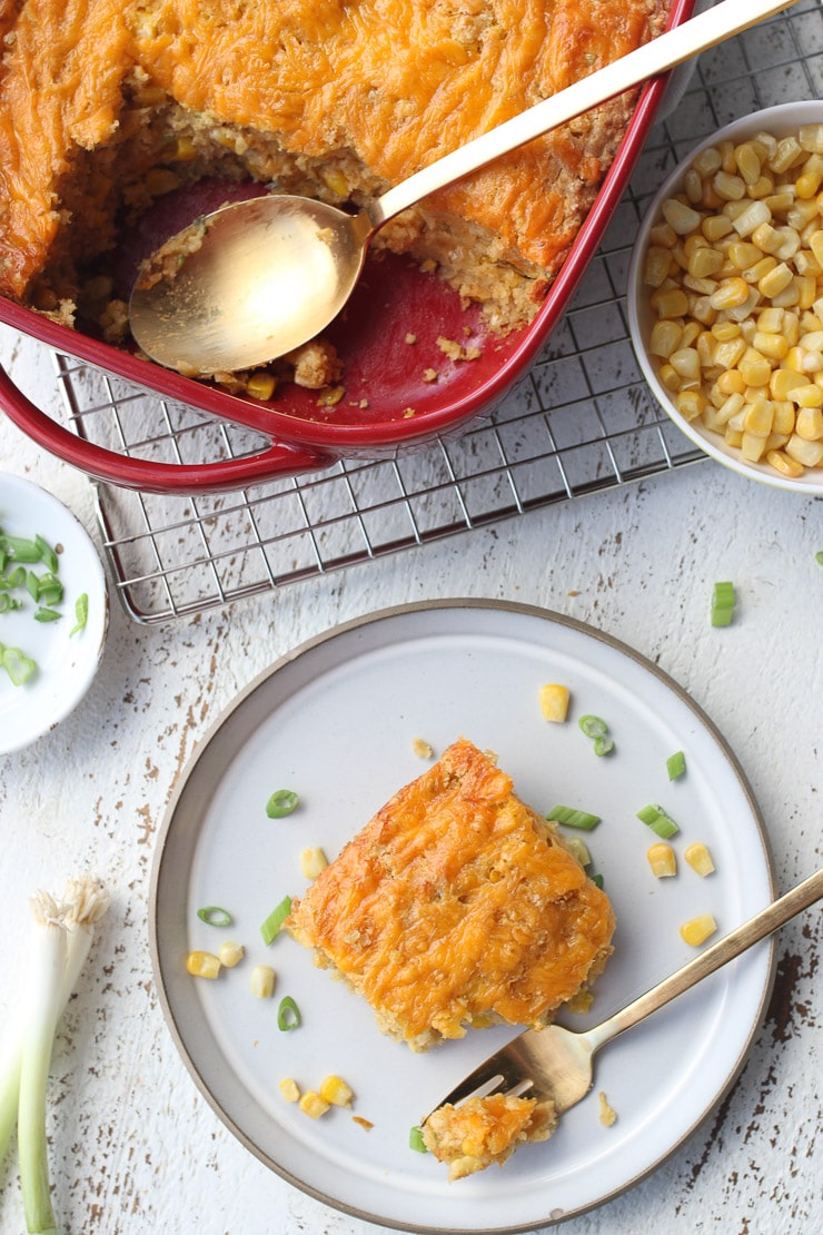 birds eye view of a white plate containing a piece of vegan corn spoonbread garnished with corn and green onions with a silver fork next to a red casserole dish and an additional bowl of fresh corn 