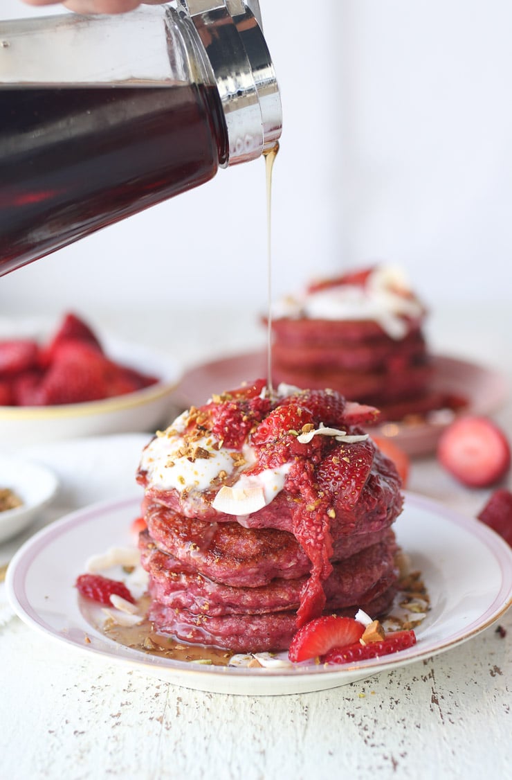 A stack of pink oatmeal pancakes topped with strawberries and drizzled with maple syrup.