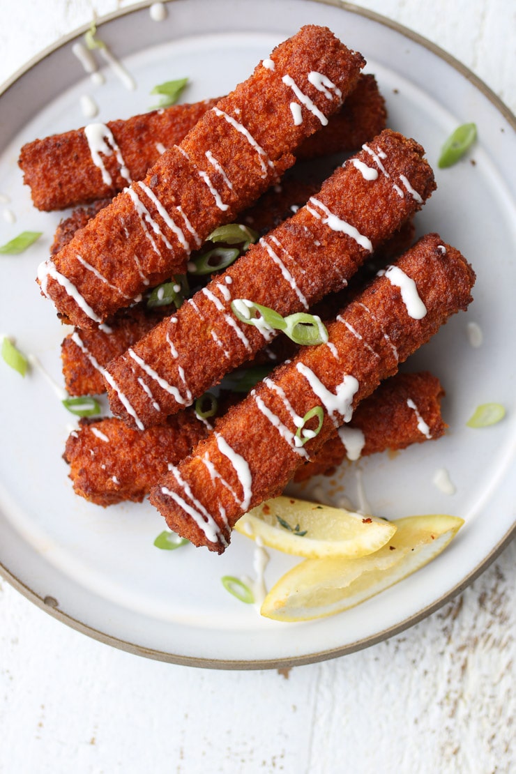 Birds eye view of tofu sticks with lemon wedges drizzled with white sauce.