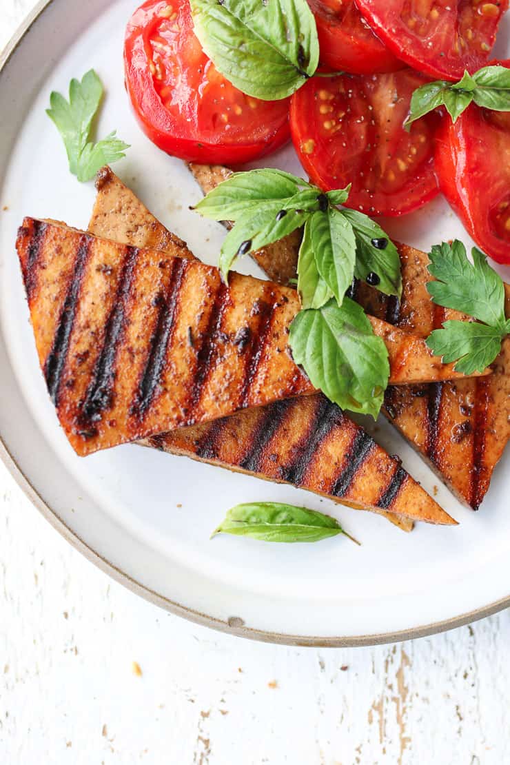 birds eye view of grilled vegan triangle steaks on a white plate with a side of tomato topped with fresh basil 