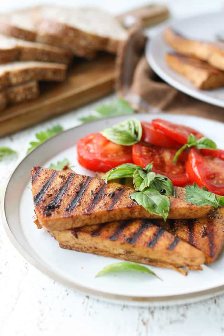 marinated grilled tofu on a white plate with tomatoes and basil on the side 