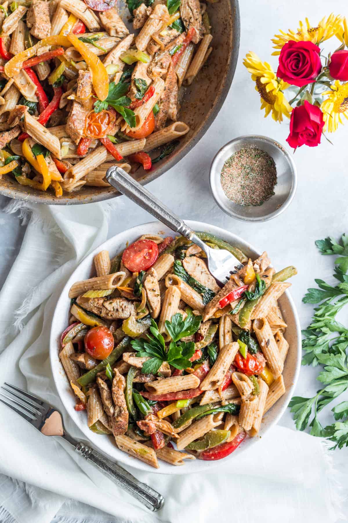 Birds eye view of jerk chicken pasta in white bowl with parsley, flowers, jerk seasoning, and a larger pot of jerk chicken pasta in the background. 