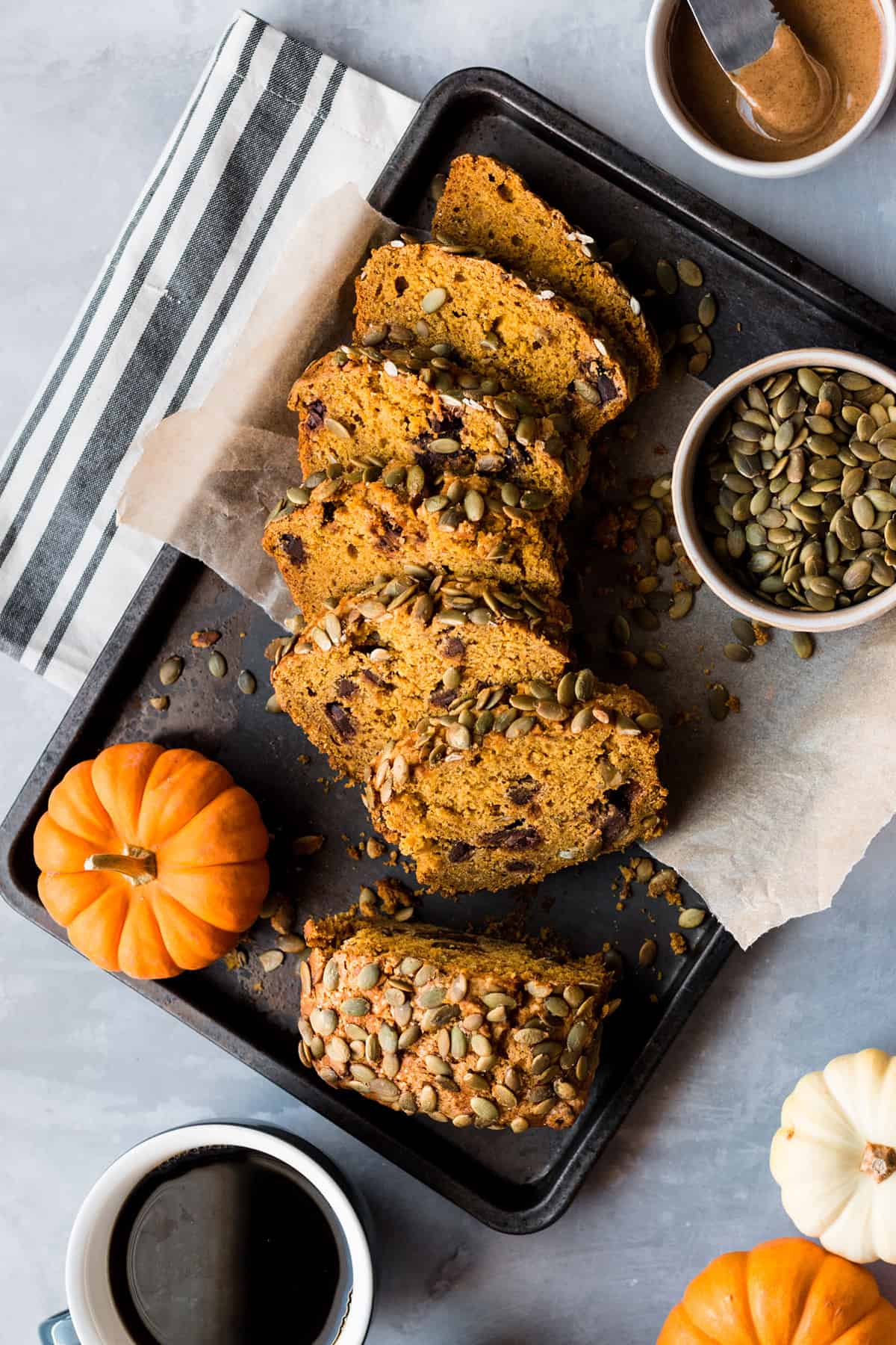 Birds eye view of vegan pumpkin bread cut into slices on a baking tray with surrounding pumpkin seeds, coffee, peanut butter, and pumpkin ornaments.