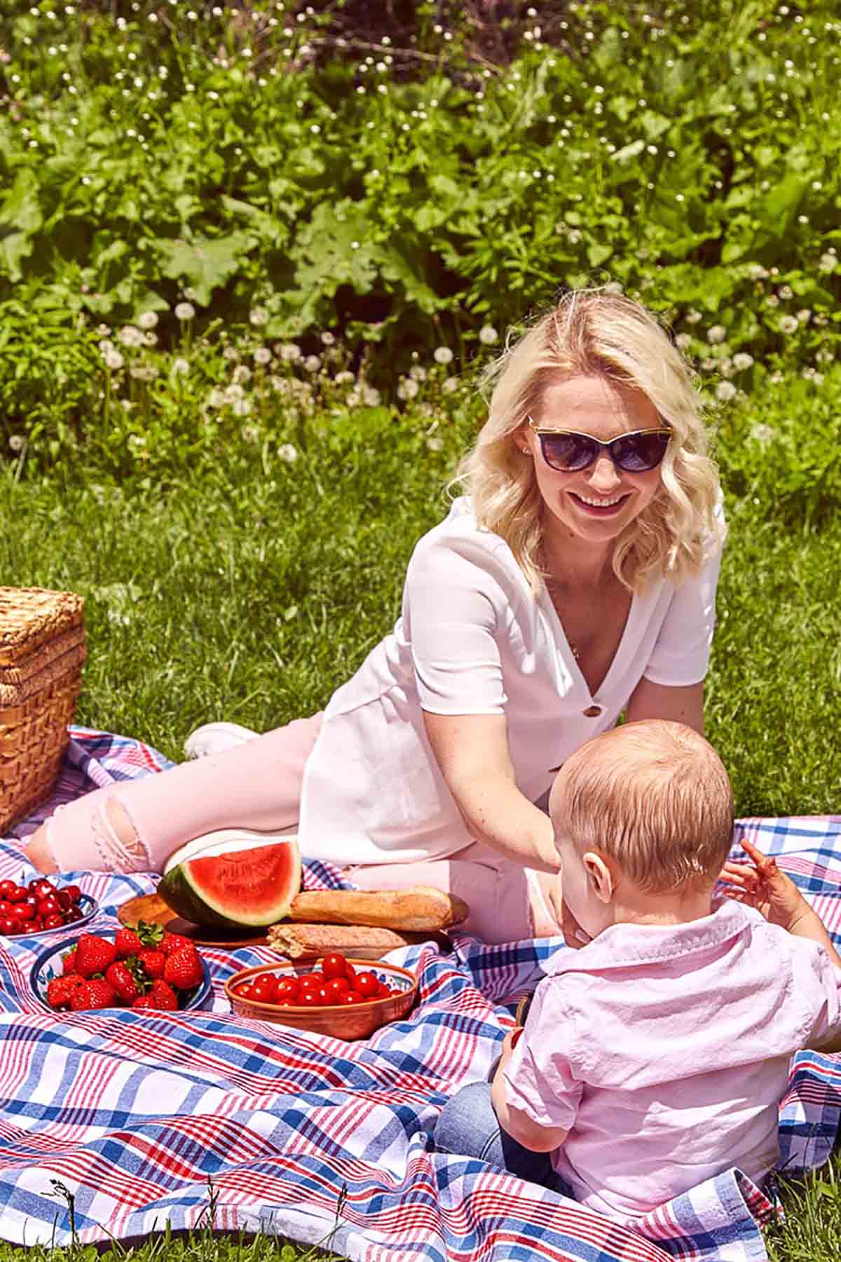 Abbey and baby E sitting a park on a picnic blanket surrounded by BLW foods like watermelon, strawberries, tomatoes, cherries, and a baguette.
