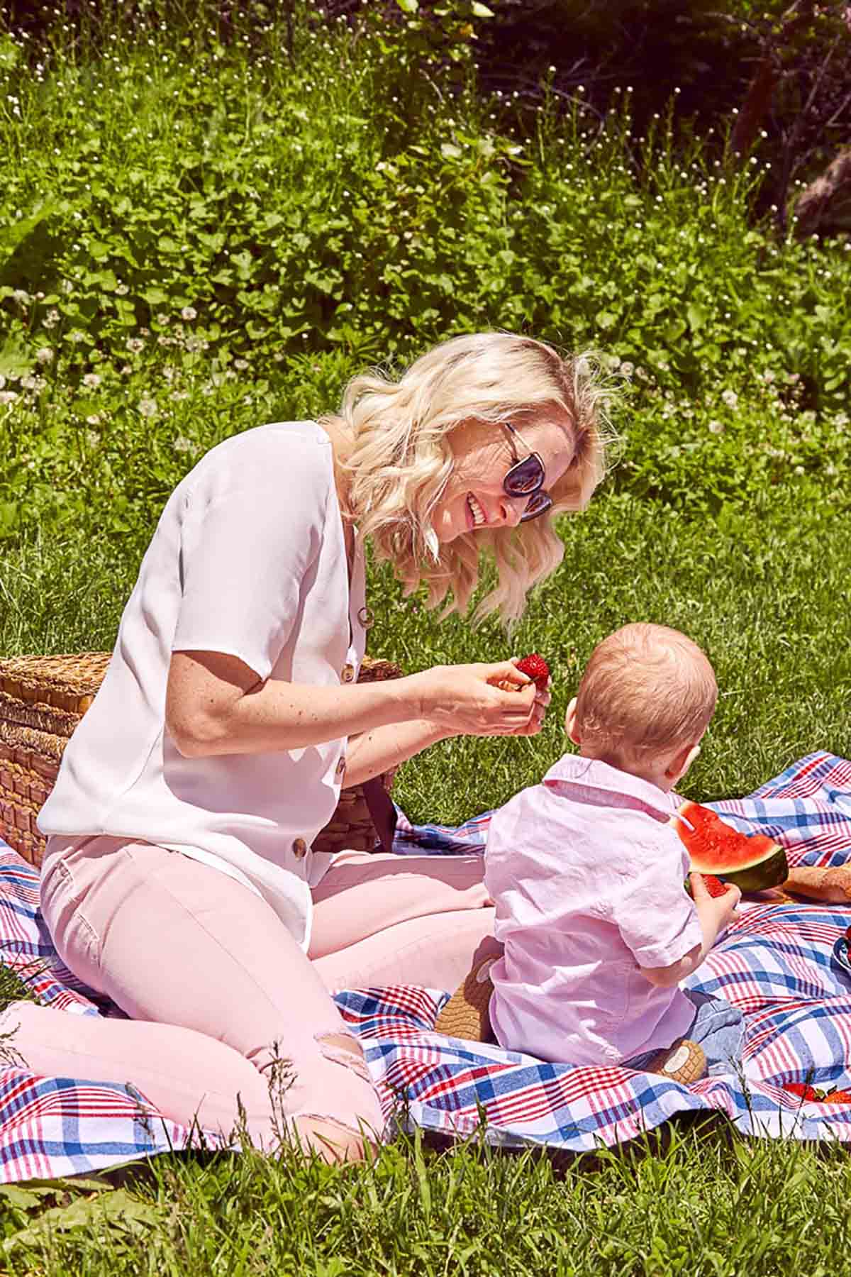 Abbey and Baby E outside sitting on a picnic blanket as Abbey feeds him a strawberry.