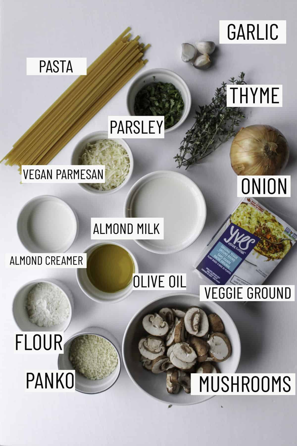 Flatlay photo of ingredients for vegan mushroom pastas: pasta, garlic, thyme, parsley, onion, vegan parmesan, almond milk, vegan milk, almond creamer, olive oil, flour, panko, and mushrooms.