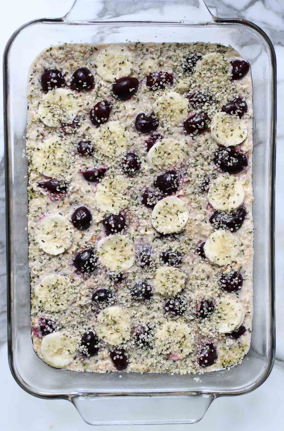 Baked oatmeal mixture added to a baking pan and topped with bananas, cherries, and hemp seeds. 