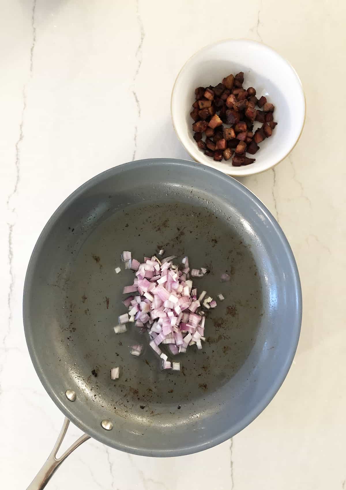 Shallot and garlic sauteed on a frying pan with pancetta on the side in a white bowl.