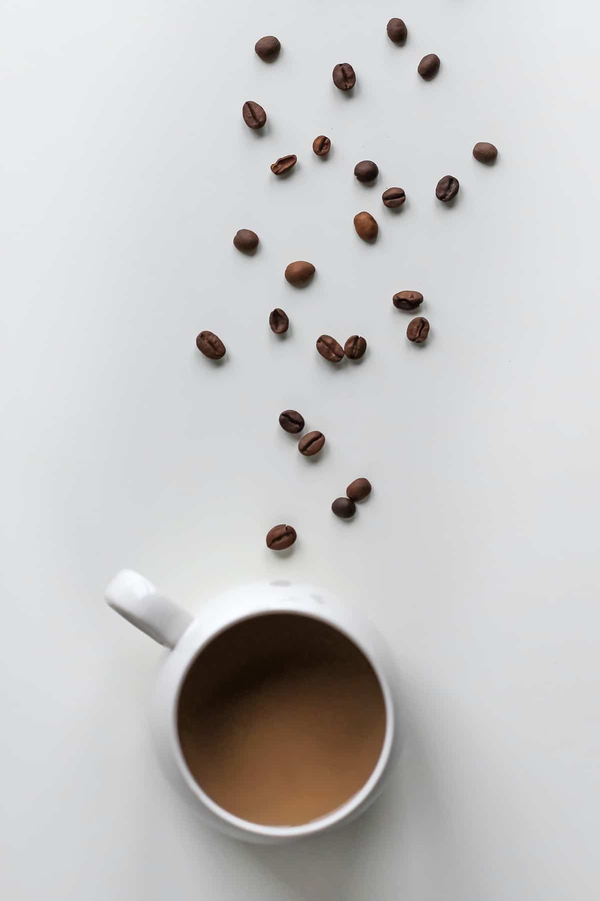 A birds eye view of coffee while intermittent fasting in a white mug with a trail of coffee beans in front of it. 