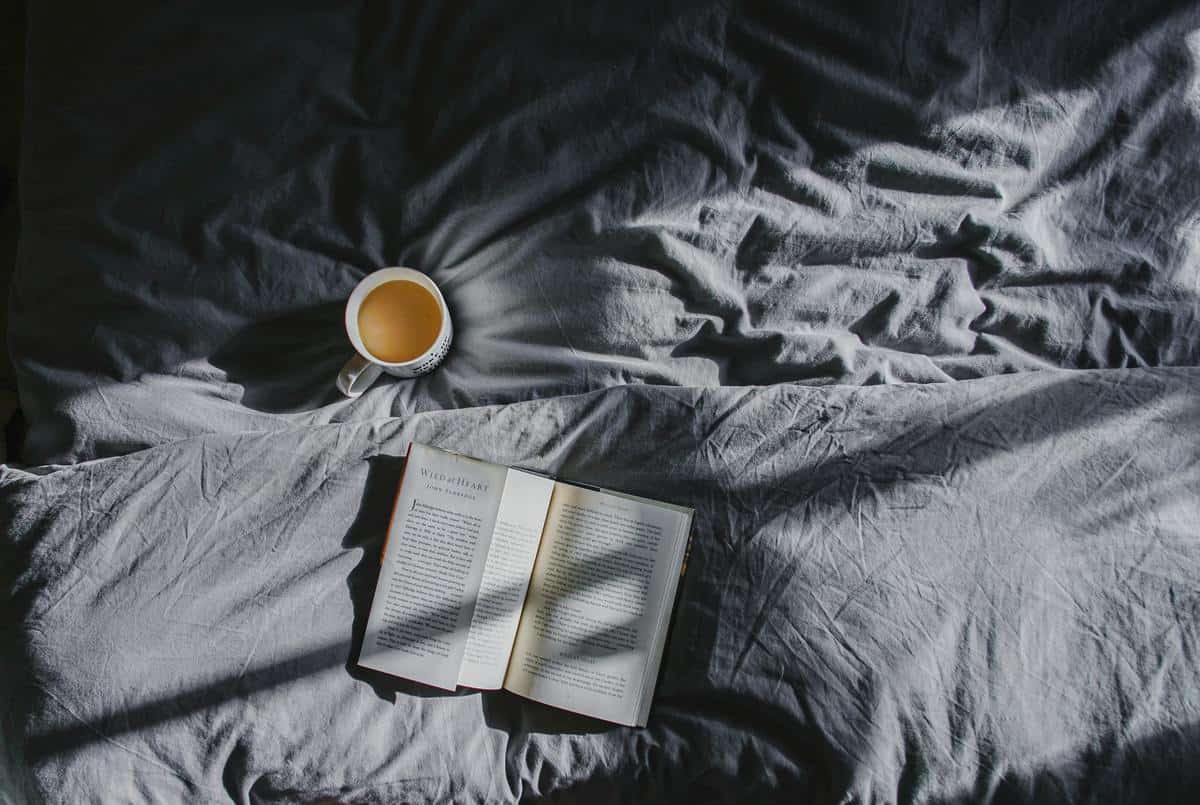 Close up image of a bed with dark grey sheets with an open book laying on a pillow, and a cup of coffee in a mug on top of the bed. 