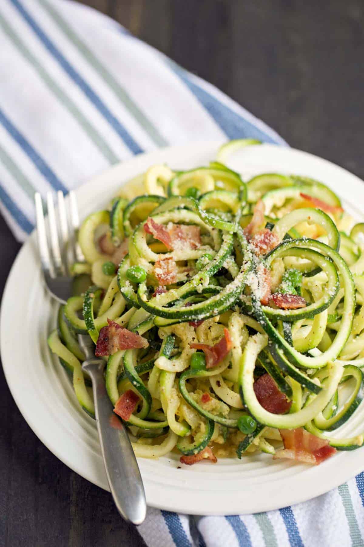 Zoodles carbonara pasta on a white plate with a fork on the side and garnished with extra parmesan cheese.