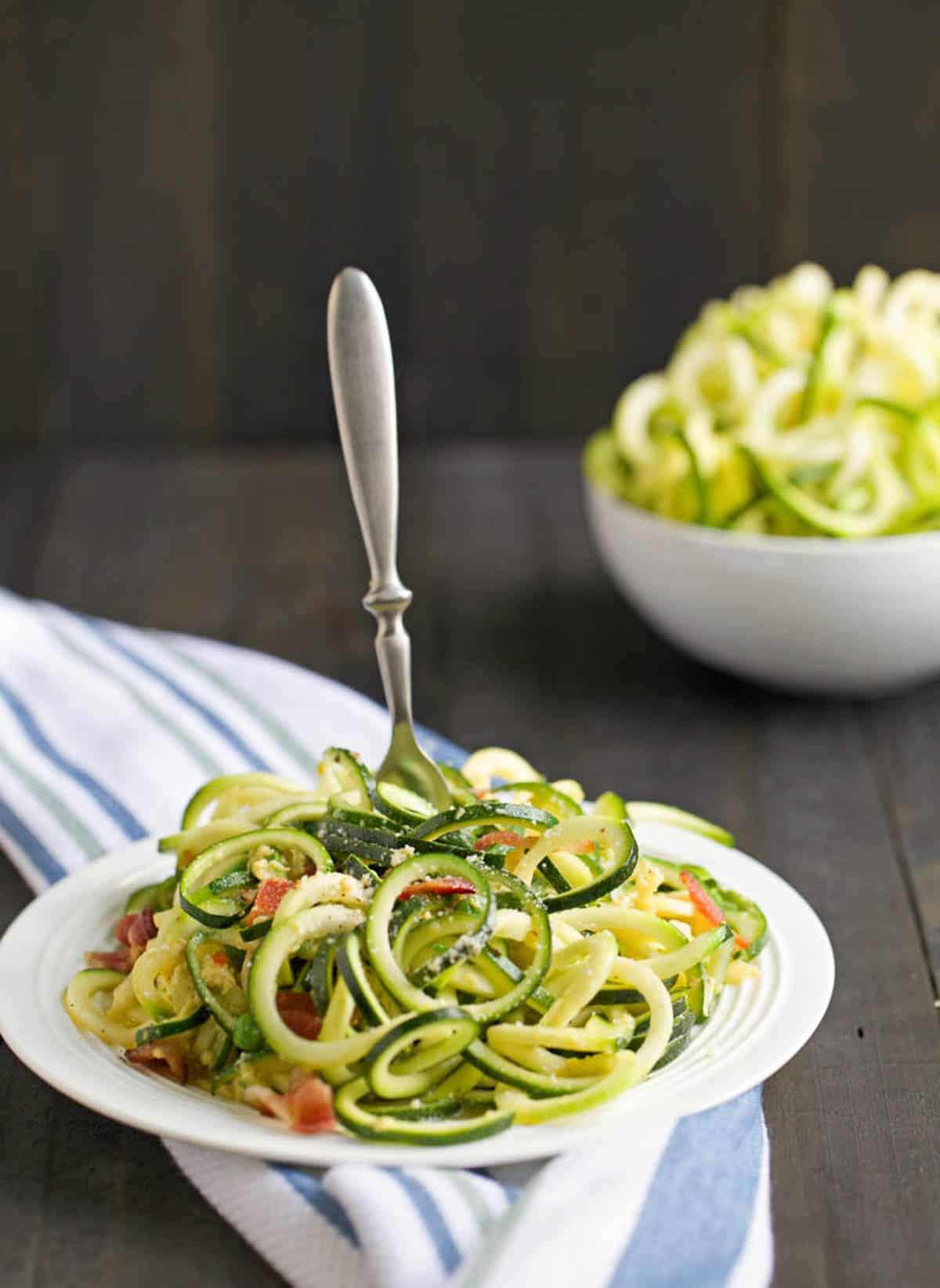 Zoodles carbonara on a white plate with a fork twisting the noodles. 