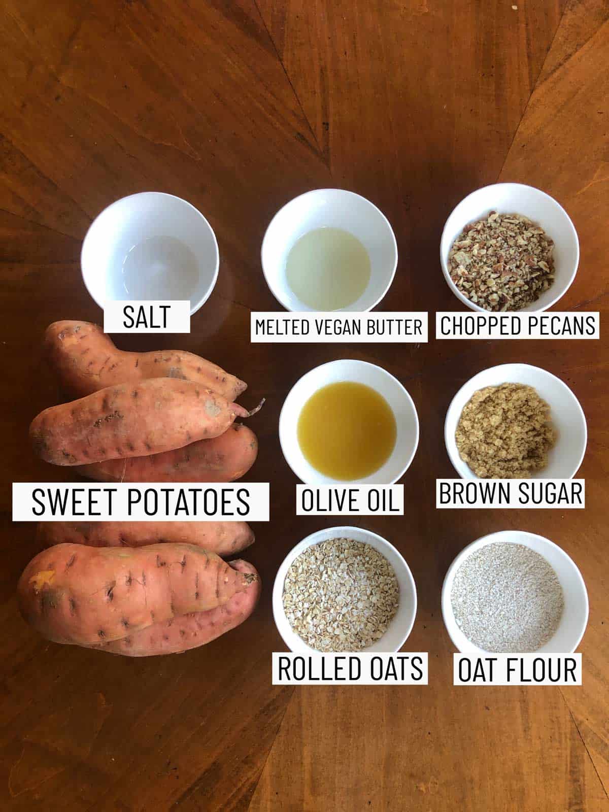 Overhead shot of ingredients to make hasselback sweet potatoes: salt, melted vegan butter, chopped pecans, sweet potatoes, olive oil, brown sugar, rolled oats, and oat flour.