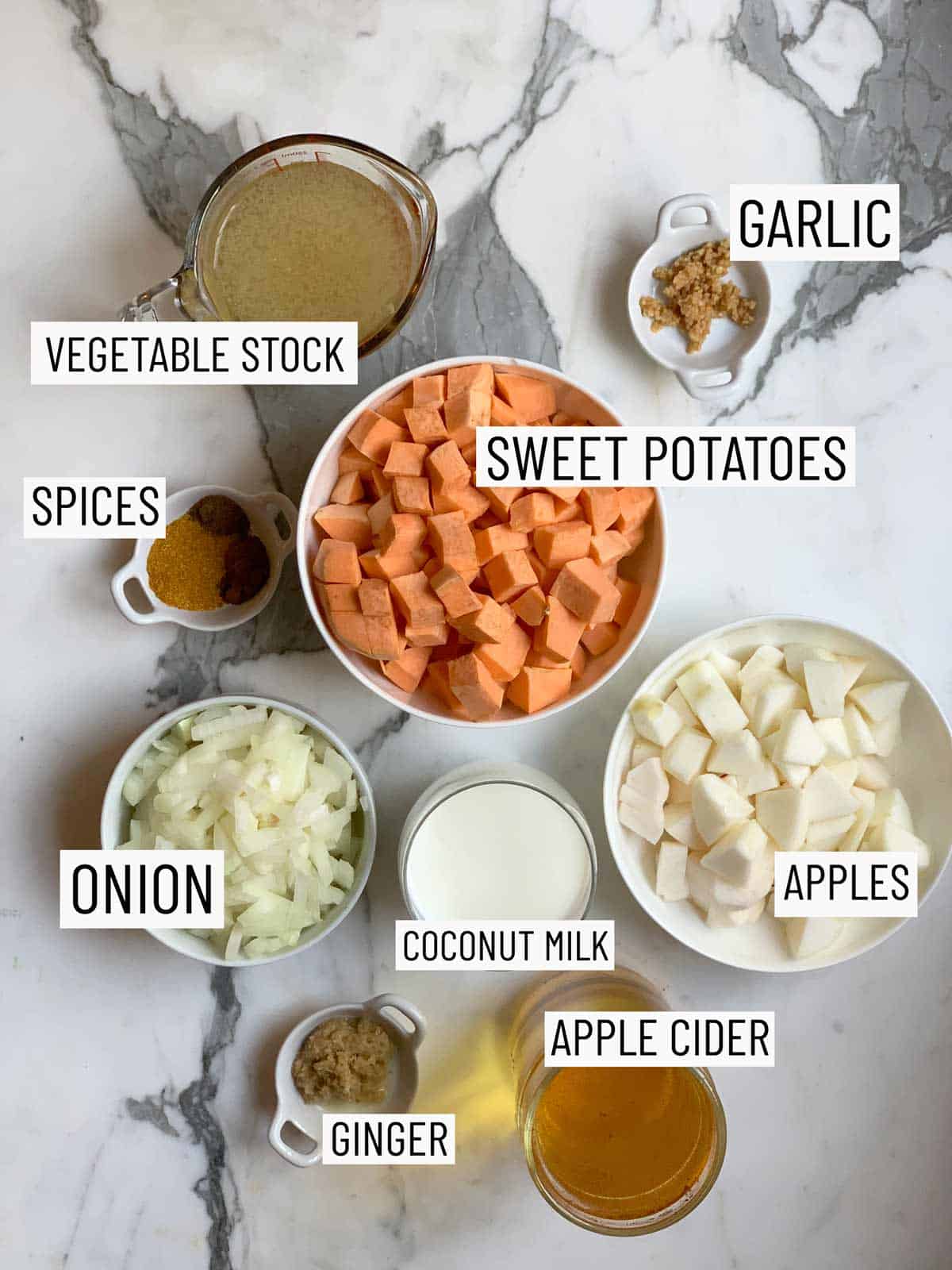 Overhead shot of recipe ingredients: vegetable stock, garlic, sweet potatoes, spices, apples, coconut milk, onion, ginger, and apple cider.