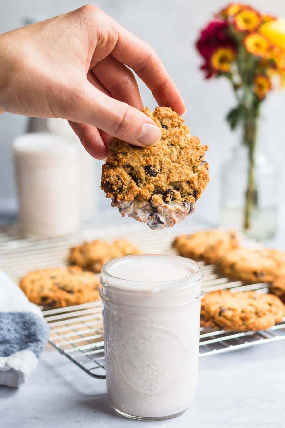 Person holding coconut flour cookie and dipping it into a glass of milk, with a baking rack with more cookies in the background. 