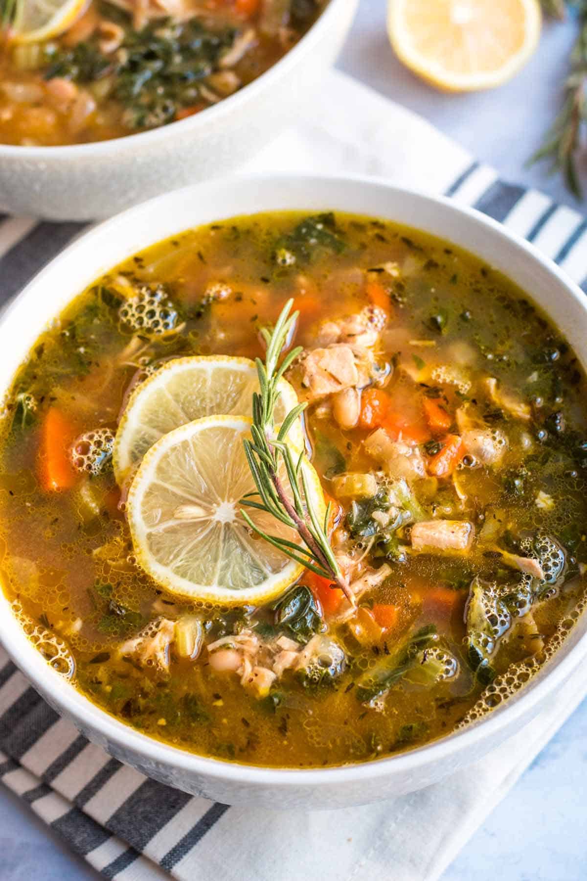 Close up of leftover turkey soup in a white bowl garnished with two lemon slices and a sprig of rosemary. 