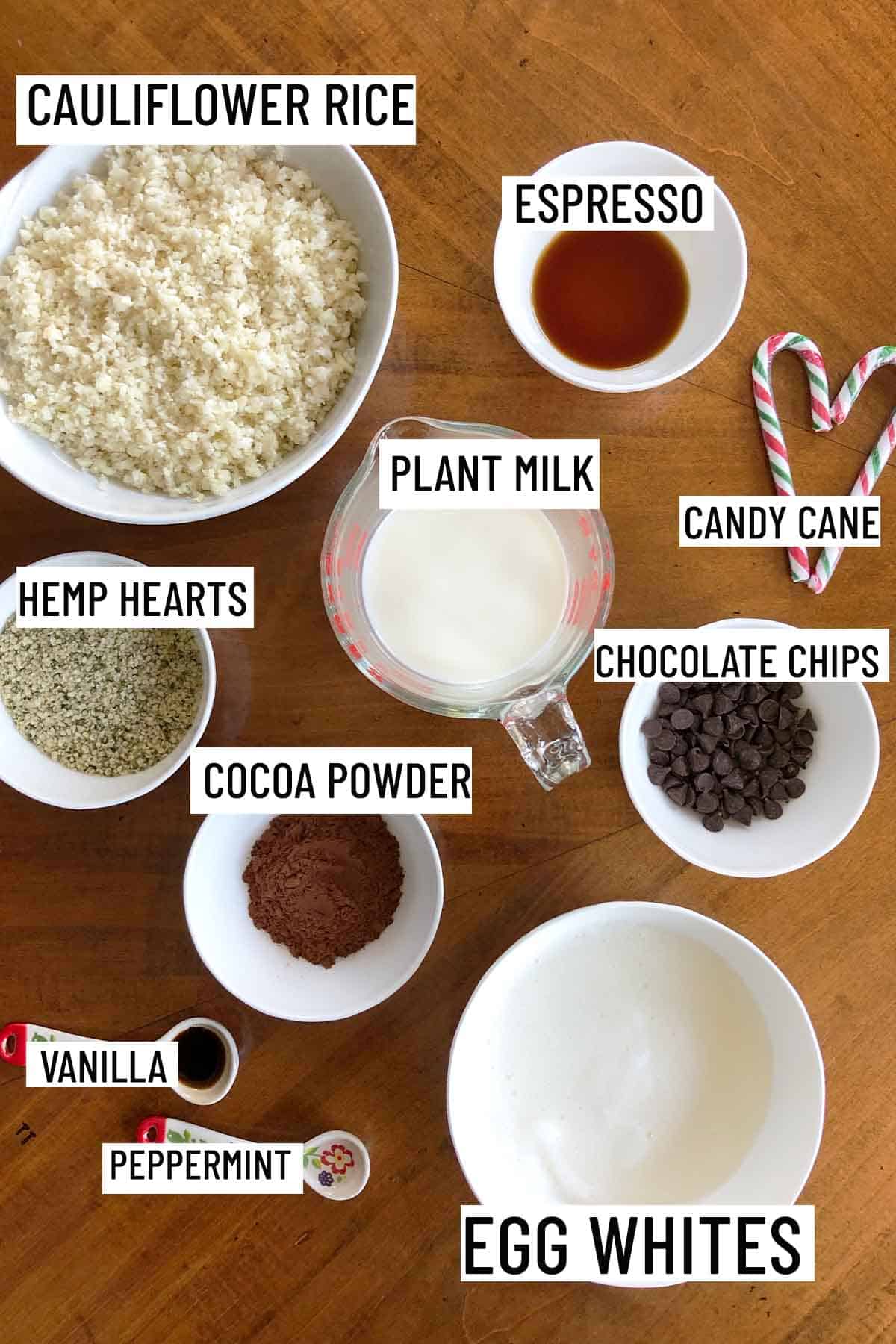 Birds eye view of portioned ingredients to make recipe including espresso, cauliflower rice, hemp hearts, plant milk, chocolate chips, candy canes, cocoa powder, egg whites, vanilla and peppermint extract.