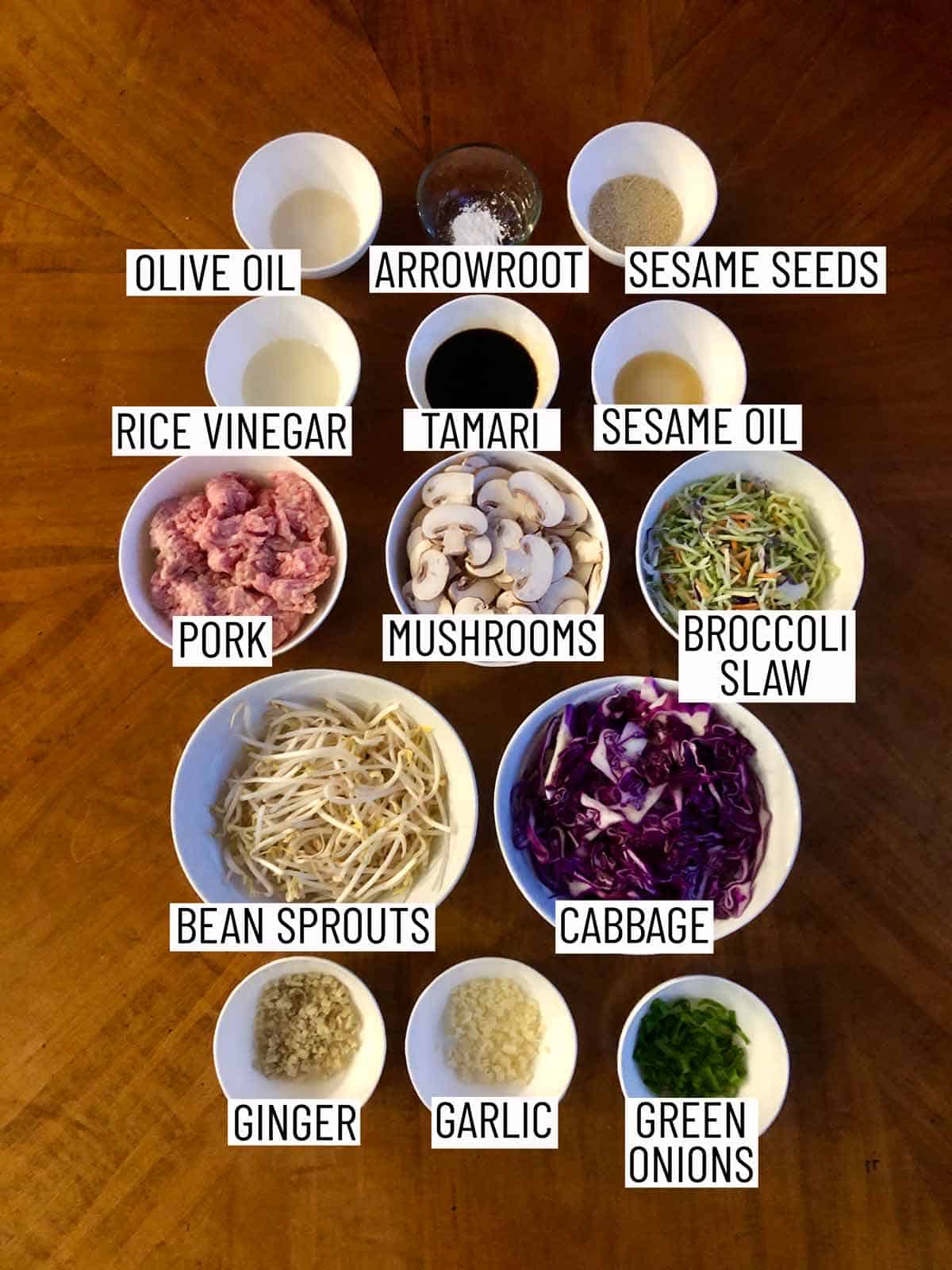 Overhead image of ingredients for egg roll in a bowl: olive oil, arrowroot, sesame seeds, rice vinegar, tamari, sesame oil, pork, mushrooms, broccoli slaw, bean sprouts, cabbage, ginger, garlic, green onions.