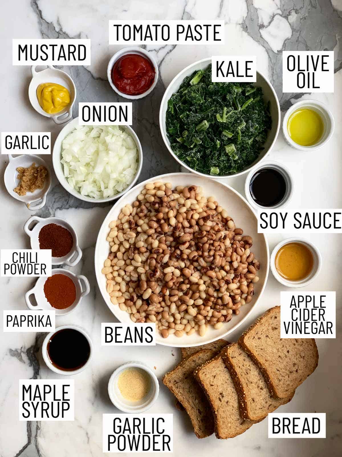 Overhead image of ingredients for beans on toast: mustard, tomato paste, olive oil, minced garlic, onion, chili powder, paprika, maple syrup, garlic powder, bread, apple cider vinegar, soy sauce, beans, and kale.