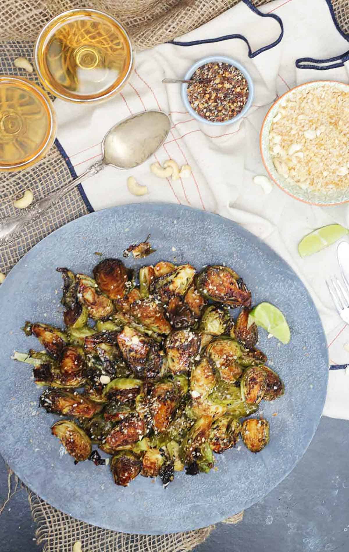 Birds eye view of spicy brussel sprouts on a blue plate with wine, cashews, and spices in the background.