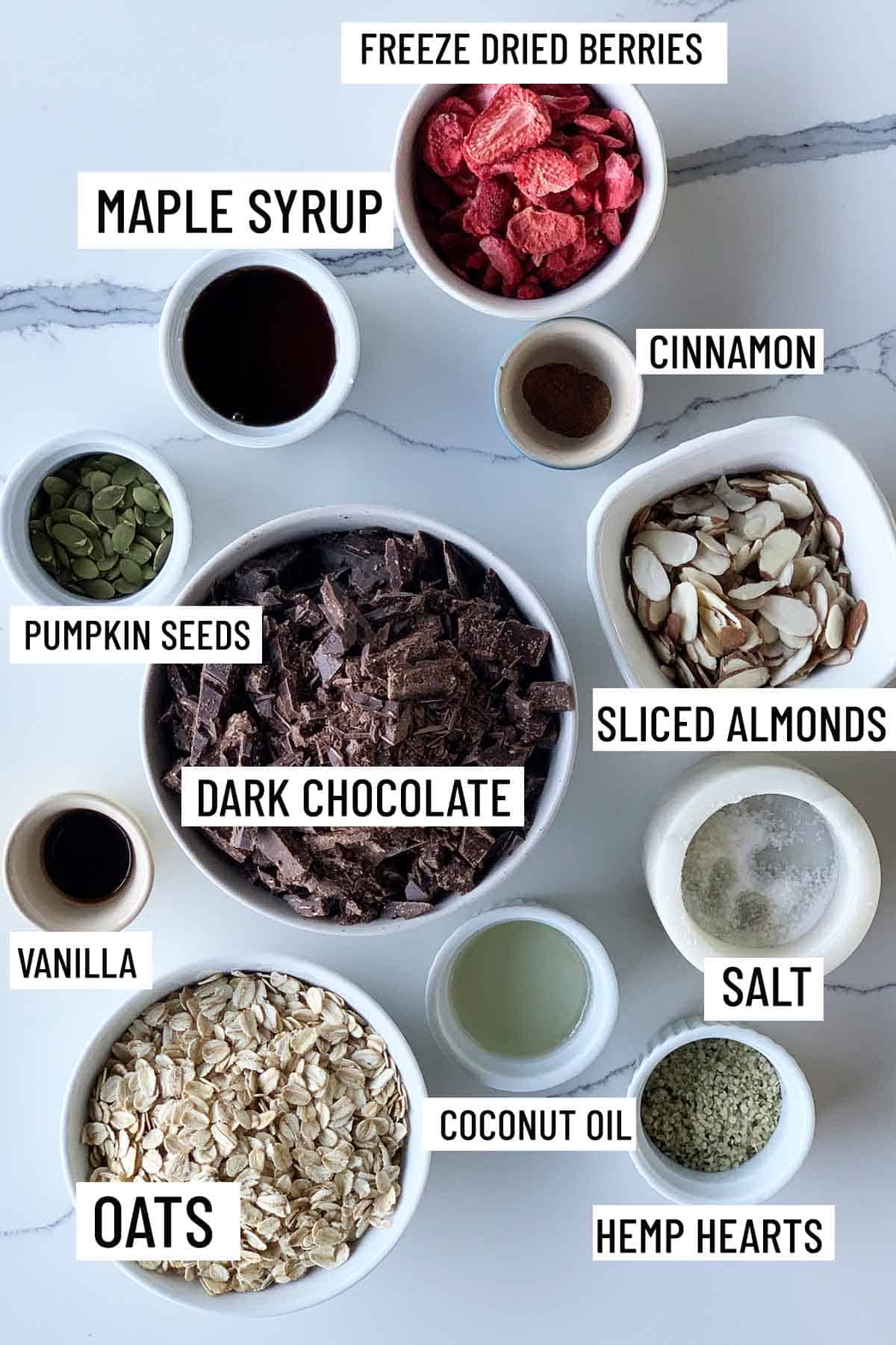 Birds eye view of portioned recipe ingredients for dark chocolate almond bark including freeze dried berries, maple syrup, cinnamon, pumpkin seeds, sliced almonds, dark chocolate, salt, vanilla, oats, coconut oil, and hemp hearts. 