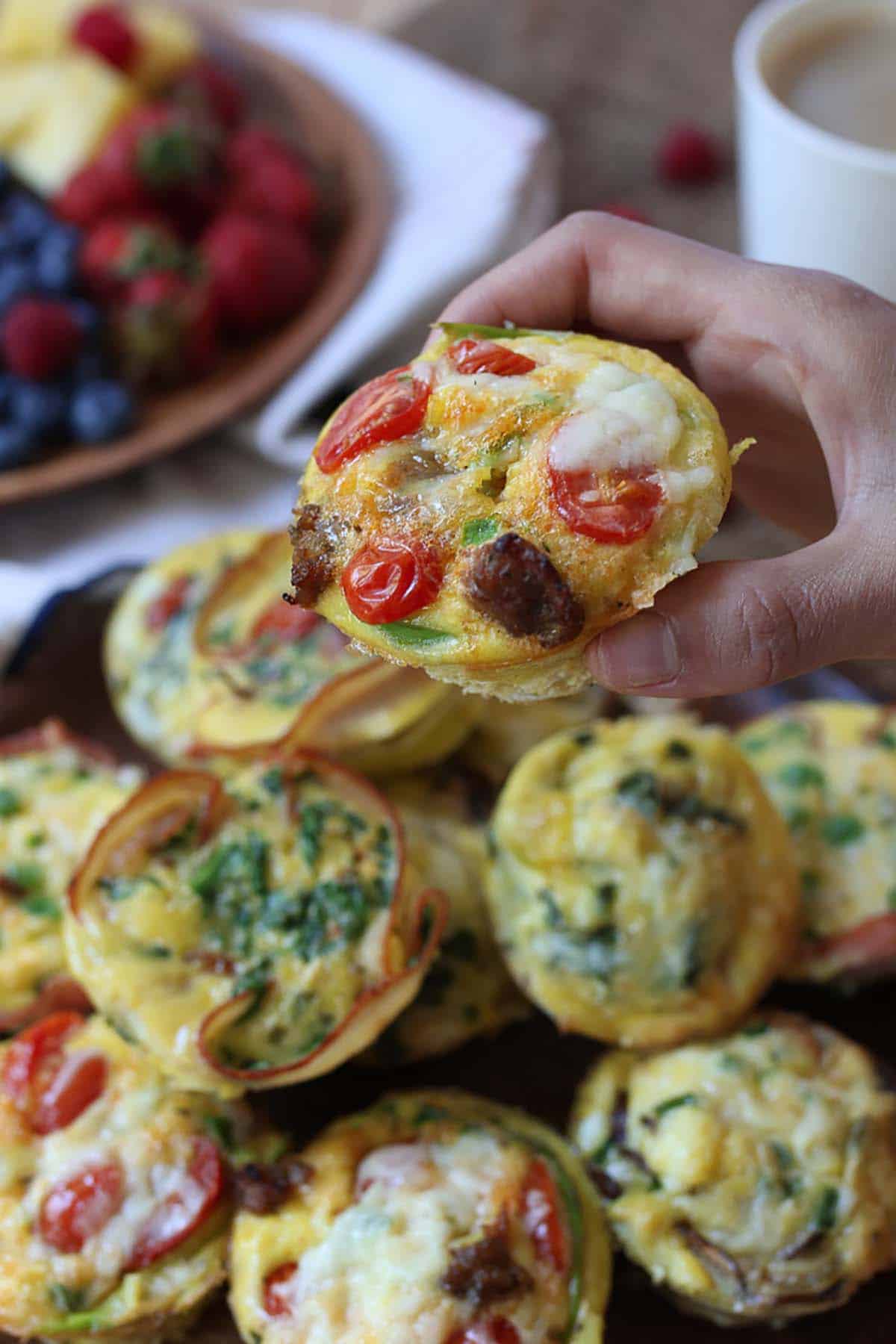 A hand picking up an egg cup with tomatoes and cheese, with a plate of egg cups in the background. 