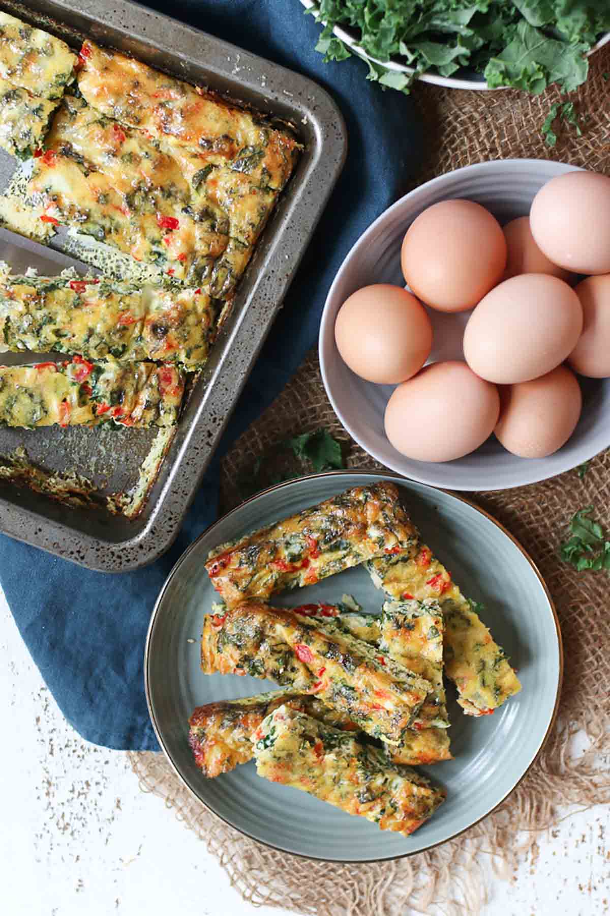 Birds eye view of a frittata cut up into slices on a plate, whole eggs in a bowl, and the remaining frittata on a baking tray. 