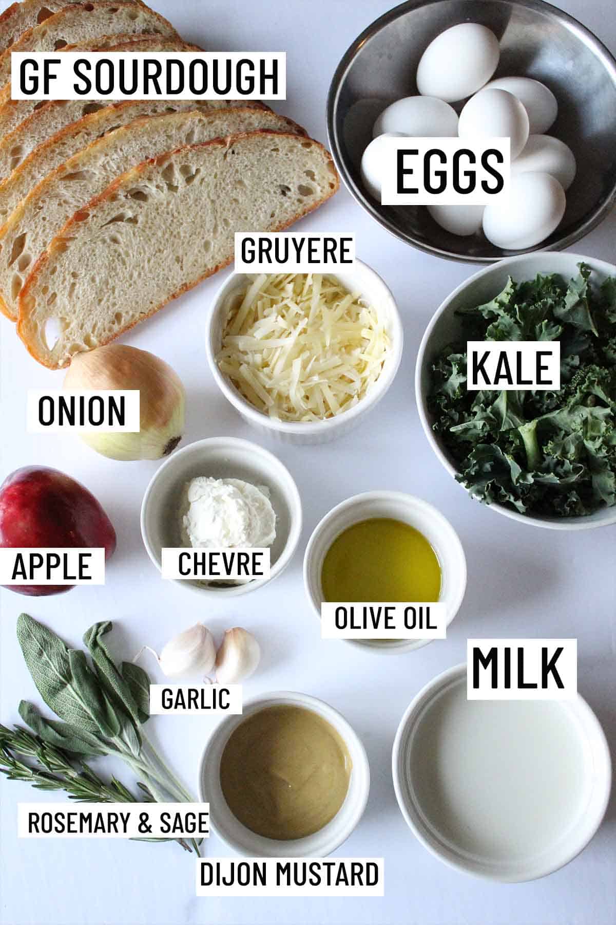 Birds eye view of strata ingredients including eggs, bread, kale, garlic, herbs, mustard, olive oil, onion, apple, and cheese. 