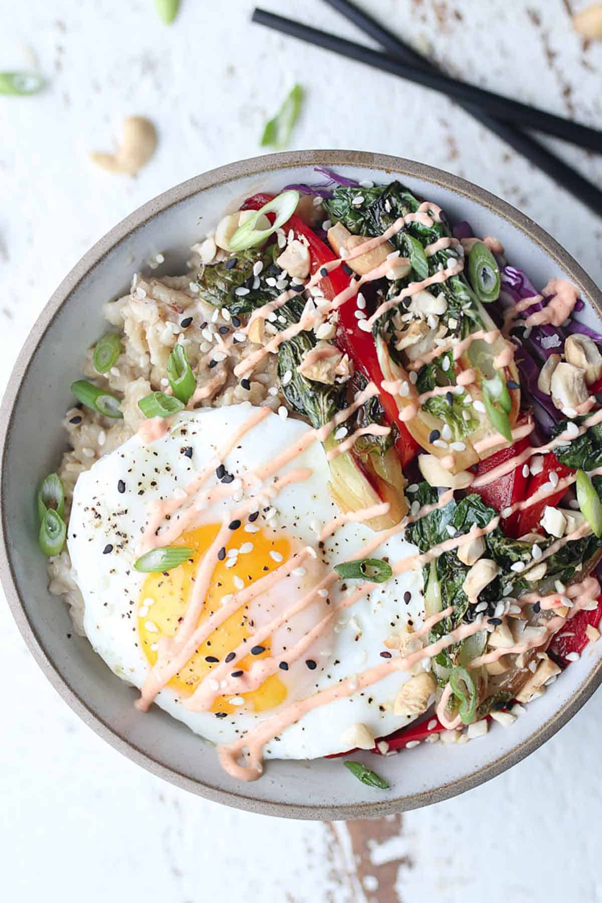 Birds eye view of a korean oatmeal bowl with an egg on top and sauteed veggies on the side and dirzzled with sauce. 