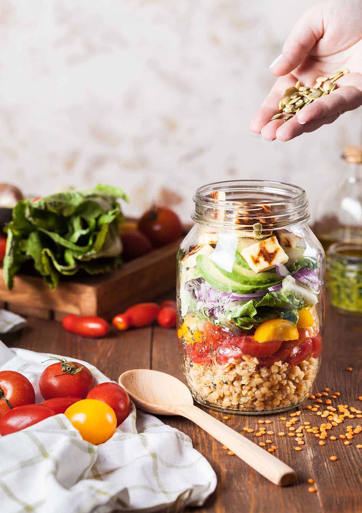 Several salad ingredients in a jar with a hand sprinkling some seeds on top. 