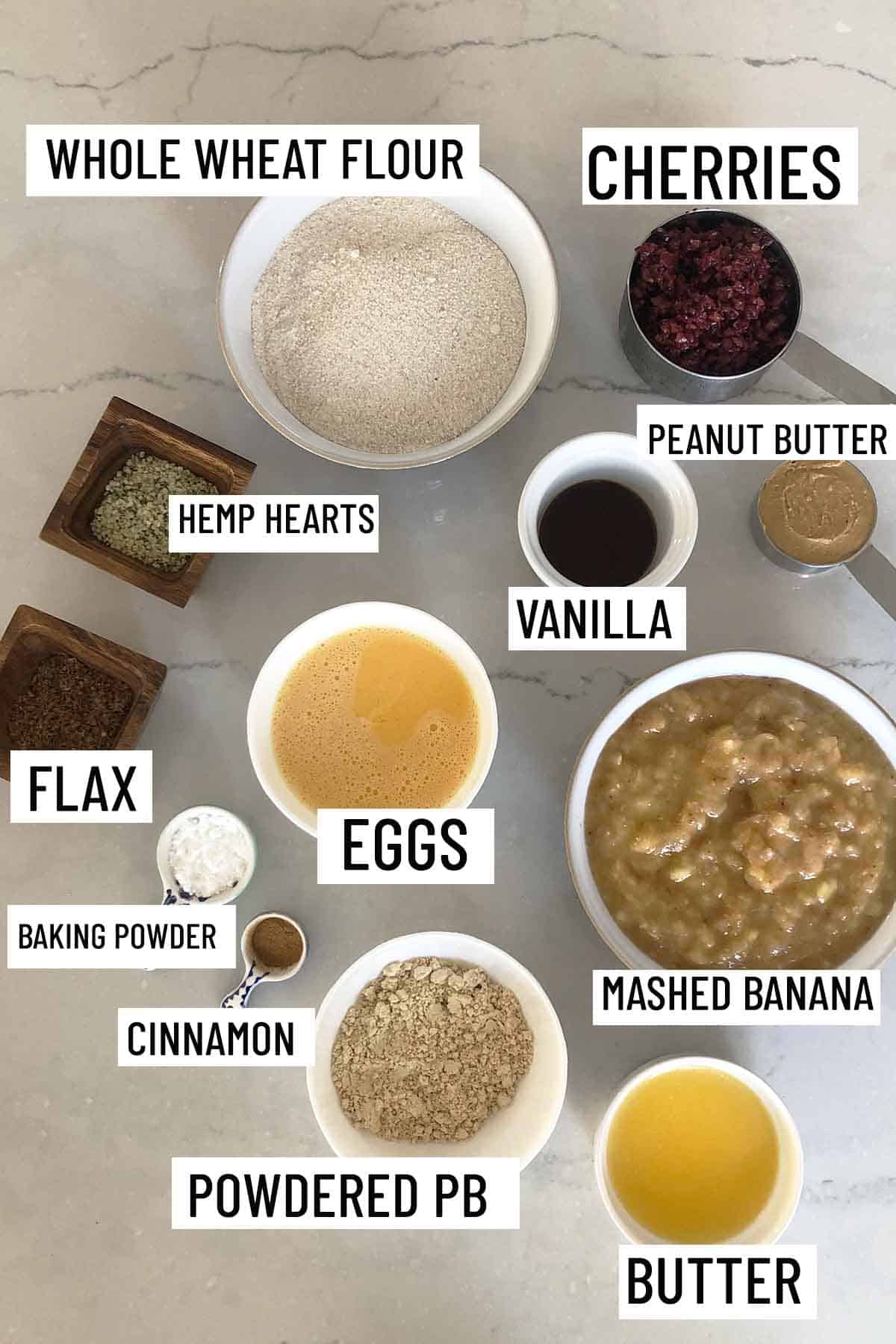 Birds eye view of portioned ingredients for recipes including butter, peanut butter, banana, cinnamon, baking powder, eggs, flax, hemp hearts, vanilla, cherries, and whole wheat flour.