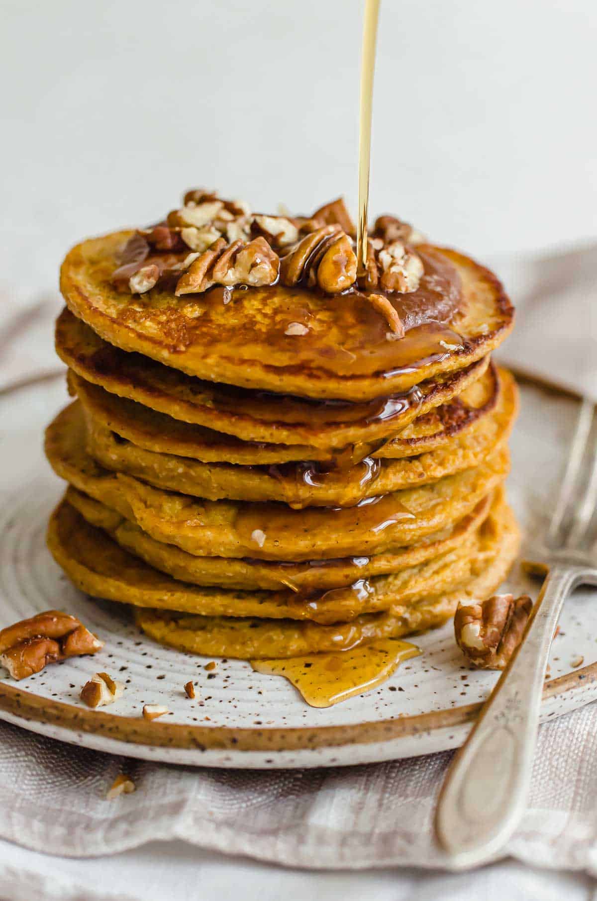 A large stack of pumpkin protein pancakes topped with pecans and being drizzled with maple syrup. 