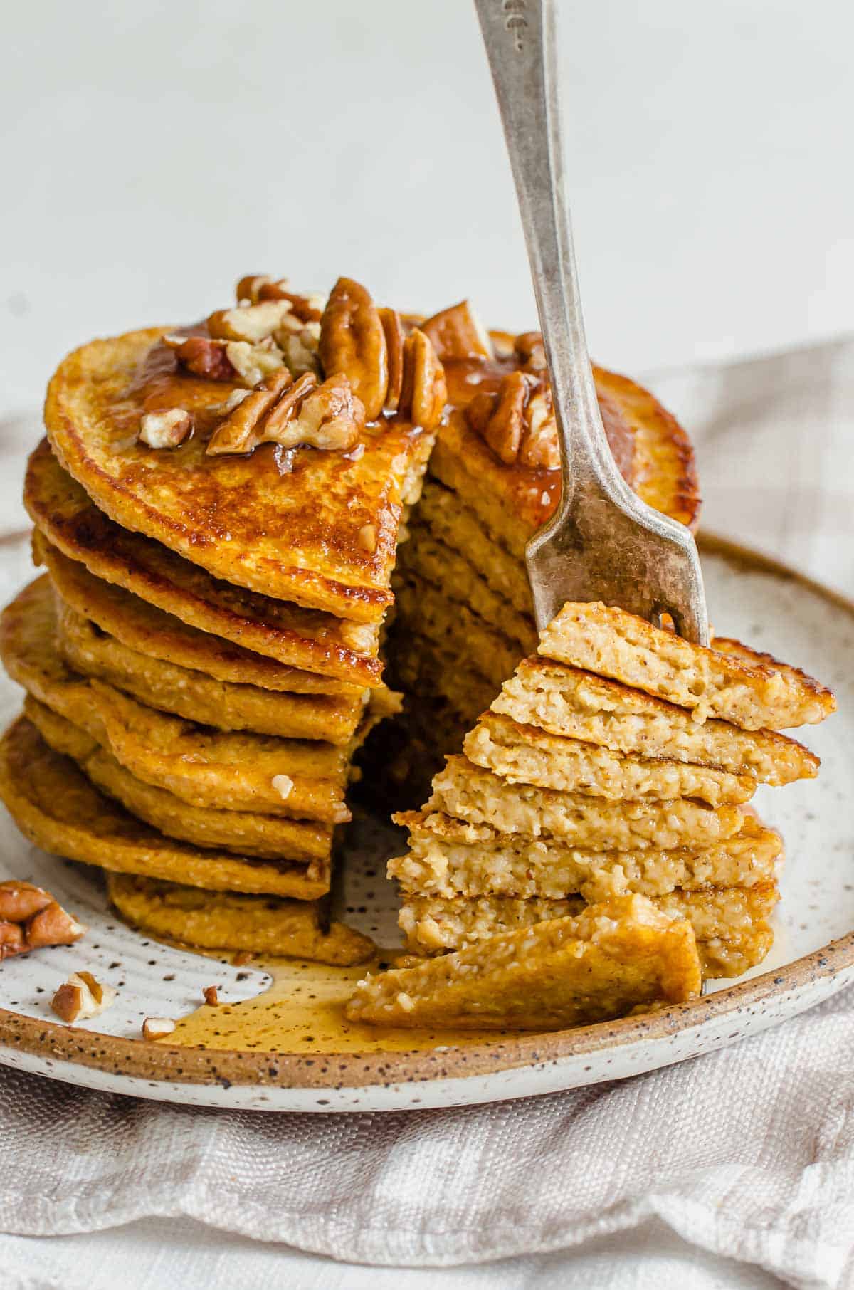 A large stack of pumpkin protein pancakes cut in the middle and topped with pecans and syrup. 