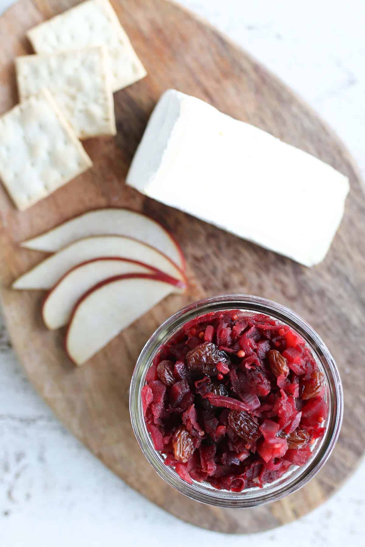 Birds eye view of apply chutney on a wooden board accompanied by cheese, slices red apple, and crackers. 