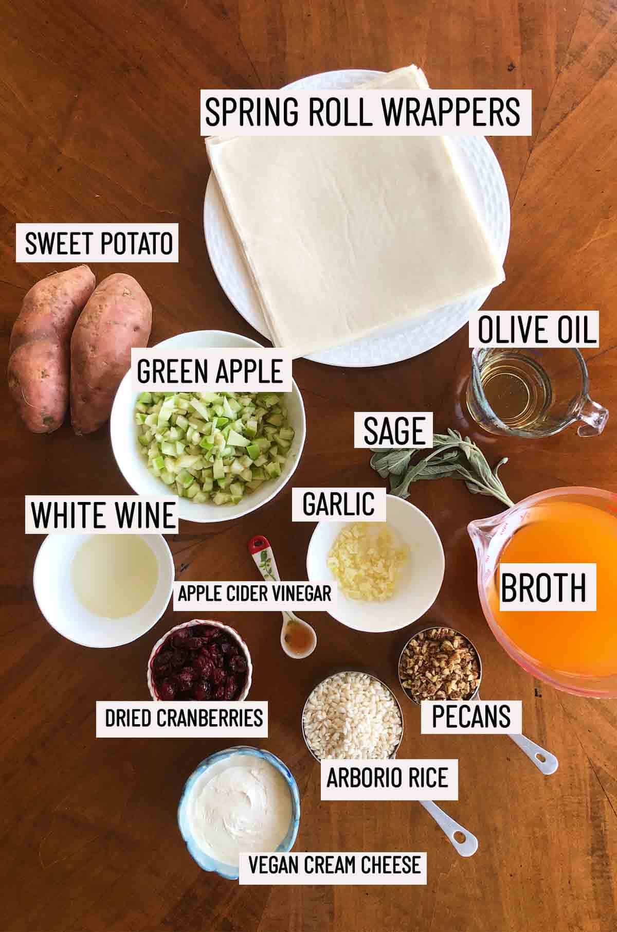 Birds eye view of portioned ingredients needed to make baked spring rolls including cream cheese, arborio rice, pecans, both, dried cranberries, apple cider vinegar, garlic, sage, olive oil, white wine, green apple, sweet potato, and spring roll wrappers. 