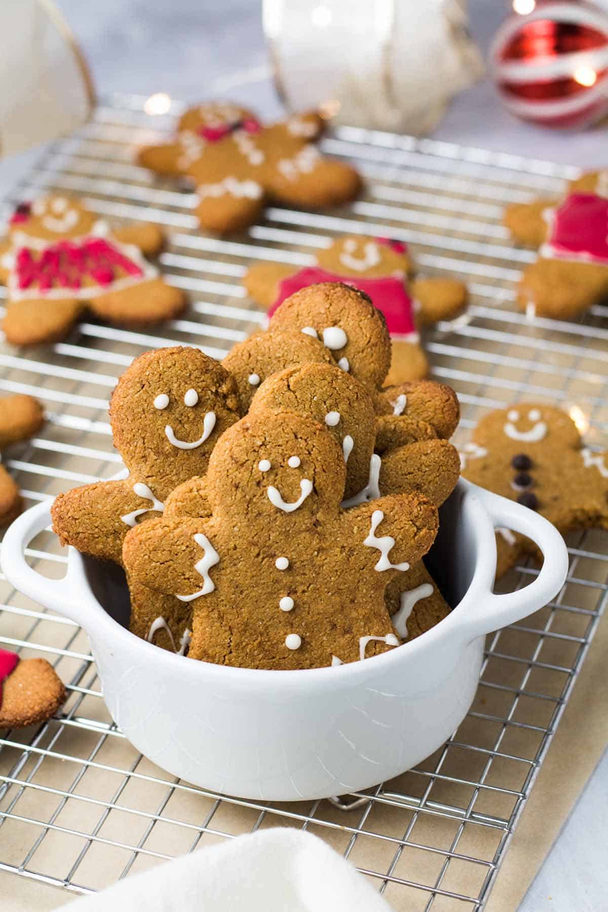 Several gingerbread cookies in a white bowl with more cookies on a baking tray in the background. 