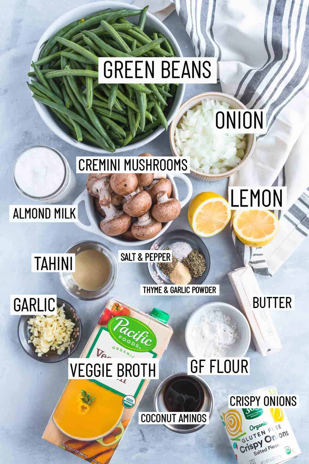 Flat lay image of portioned recipe ingredients including onion, lemon, mushroom, almond milk, green beans, tahini, spices, butter, flour, veggie broth, garlic, coconut aminos, and crispy onions. 