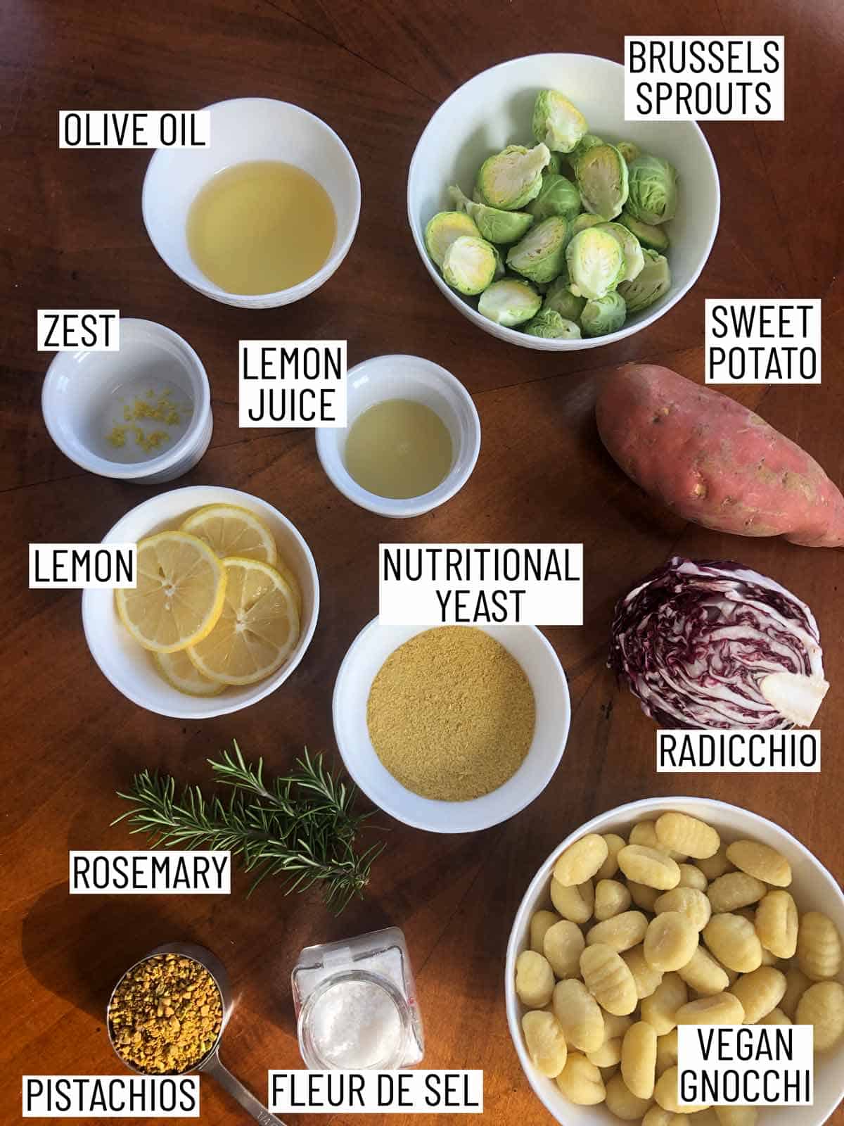 Flat lay image of portioned recipe ingredients including sweet potato, radicchio, vegan gnocchi, fleur de sel, pistachios, rosemary, lemon, nutritional yeast, olive oil, and brussels sprouts.