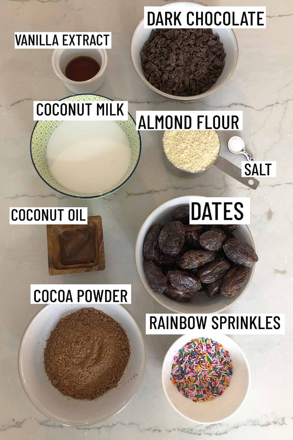Birds eye view of portioned recipe ingredients for no bake brownies including as rainbow sprinkles, coconut milk, coconut oil, cocoa powder, almond flour, salt, vanilla extract, and dark chocolate. 