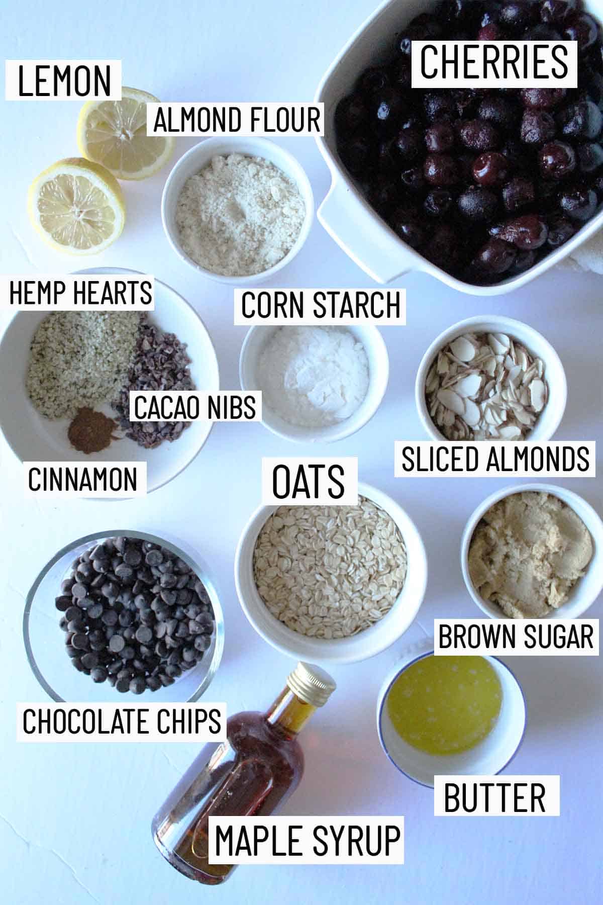 Flat lay of portioned recipe ingredients including brown sugar, butter, chocolate chips, oats, cacao nibs, cinnamon, hemp hearts, lemon, corn starch, almond flour, and cherries. 