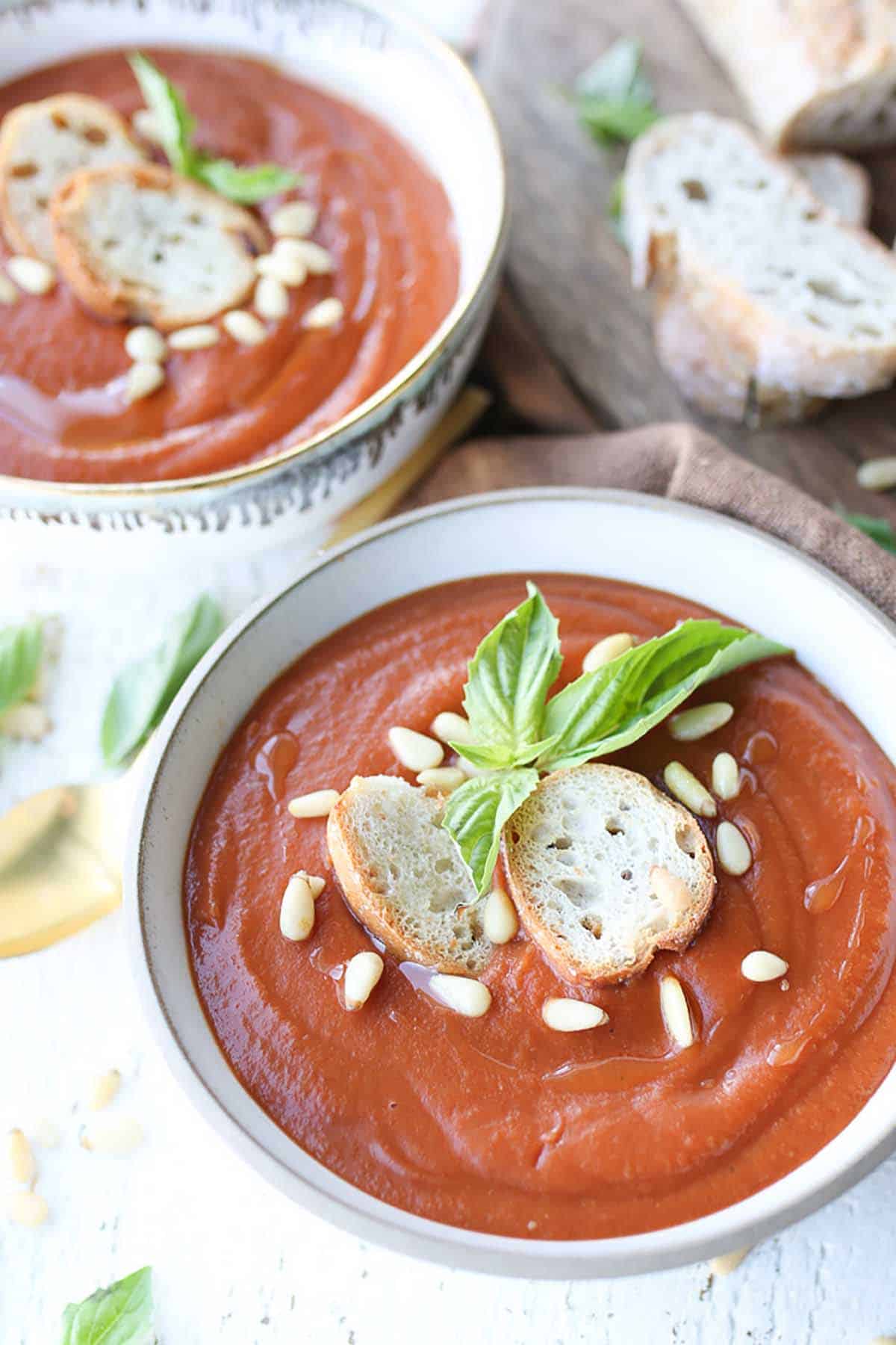 Close up of tomato soup in a white bowl topped with pine nuts, baguette slices, and basil. 