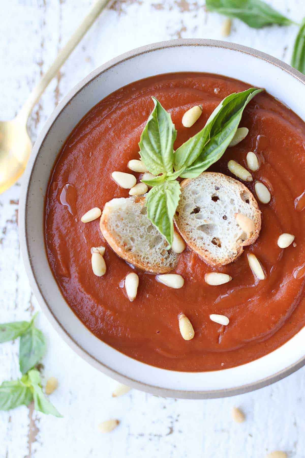Close up birds eye view image of creamy tomato soup in a white bowl topped with pin nuts, baguette slices and fresh basil. 