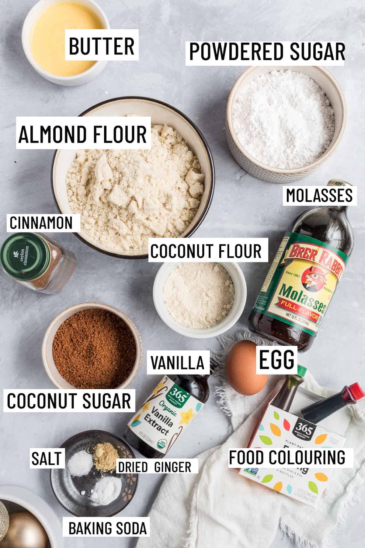 Birds eye view of portioned recipe ingredients including almond flour, icing sugar, coconut flour, coconut sugar, cinnamon, egg, molasses, food colouring, dried ginger, salt, baking soda, butter and vanilla. 