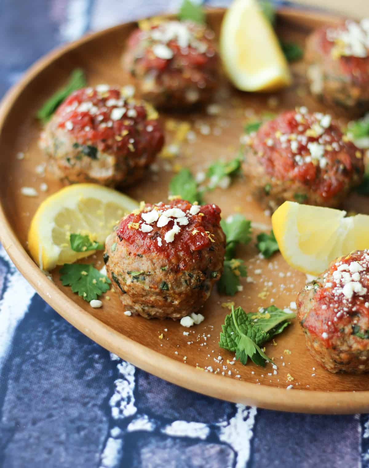 Close up of Greek meatballs topped with tomato sauce and feta, garnished with mint and lemon wedges. 