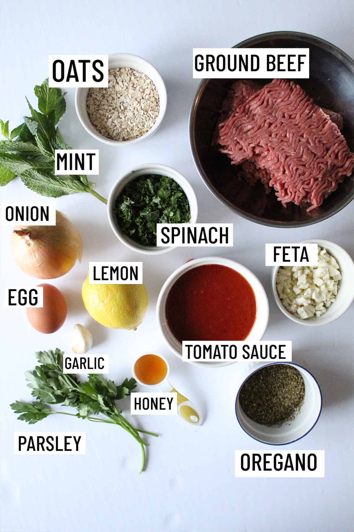 Birds eye view of portioned recipe ingredients for meatloaf muffins including ground beef, lemon, onion, garlic, egg, oregano, mint, parsley, oats, honey, feta, spinach and tomato sauce.