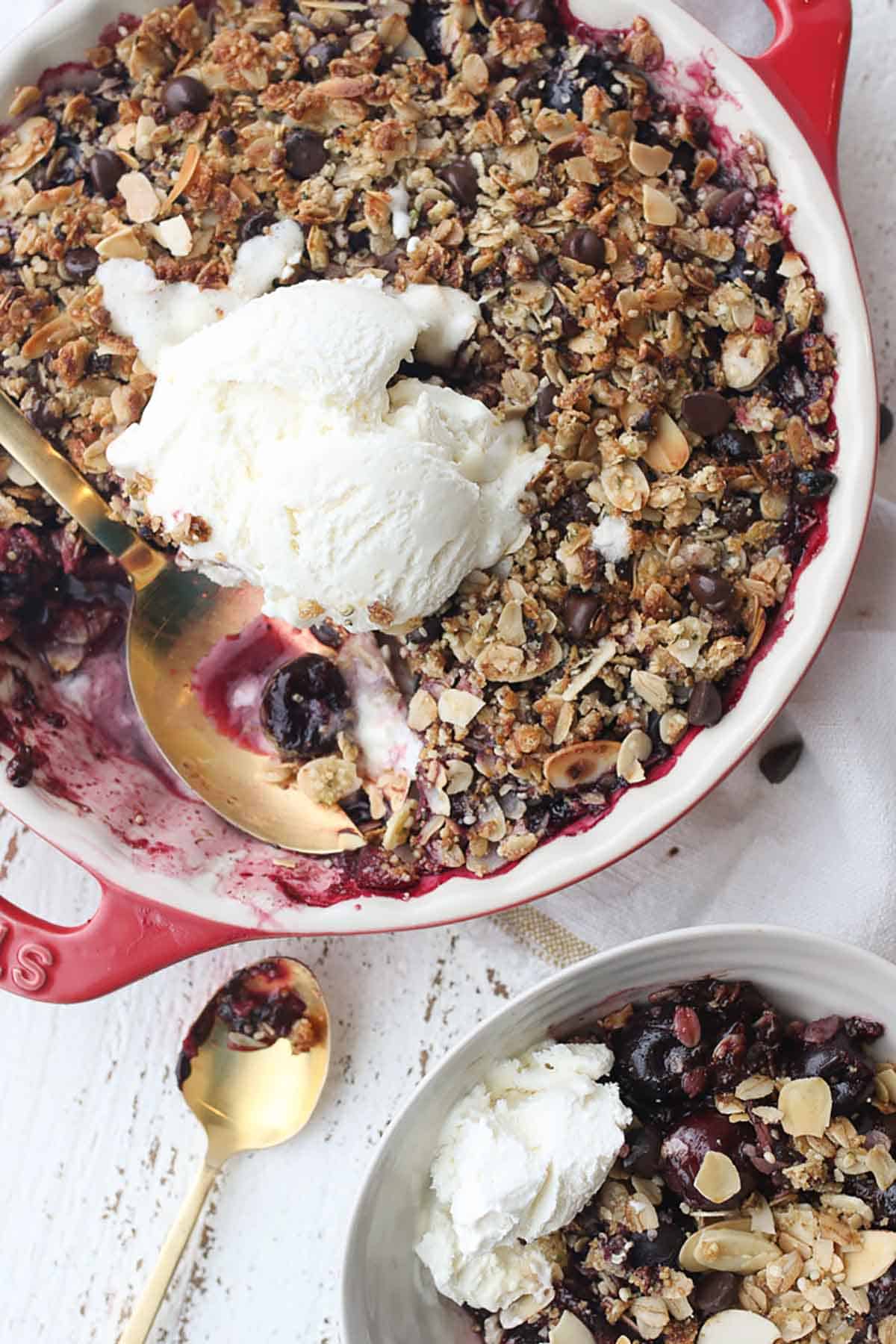 Birds eye view of chocolate cherry crisp in a round red baking dish topped with ice cream, with a serving bowl of crisp on the side. 
