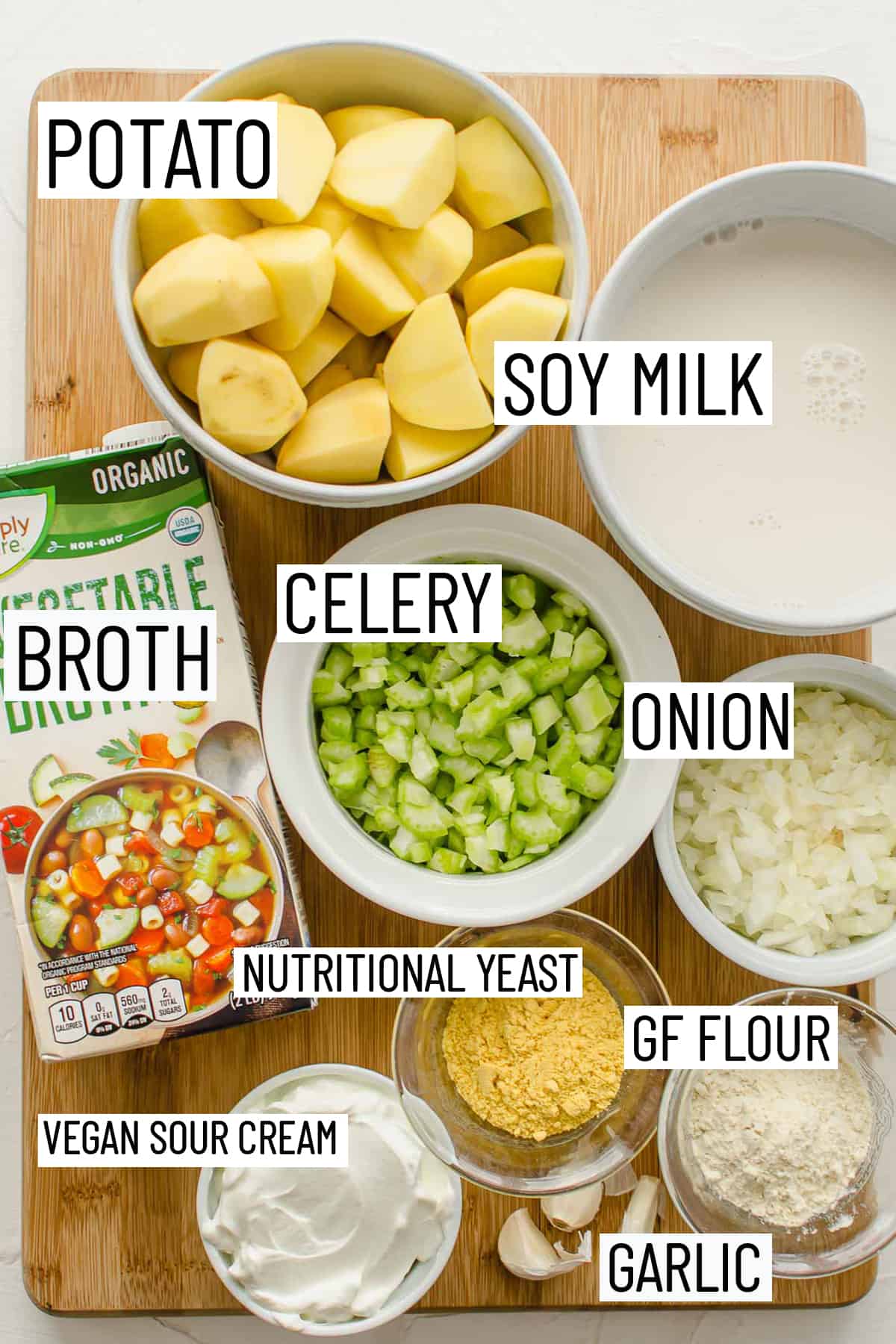 Flat lay image of portioned ingredients for potato soup including nutritional yeast, flour, sour cream, garlic, onion, celery, broth, soy milk, and potato. 